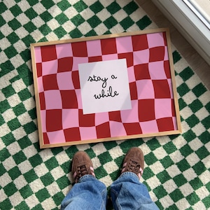 Könnte beinhalten: Ein gerahmtes Kunstwerk mit einem rosa-roten Schachbrettmuster. Ein weißes Quadrat mit den Worten "stay a while" ist auf dem Kunstwerk zentriert. Das Kunstwerk liegt auf einem grün-weißen Schachbrettteppich.