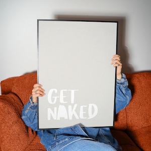 Könnte beinhalten: Ein gerahmtes Poster mit den Worten "GET NAKED" in weißer Schrift. Das Poster wird von einer Person gehalten, die eine Jeansjacke und Jeans trägt und auf einem orangefarbenen Sofa sitzt. Das Poster hat einen schwarzen Rahmen.