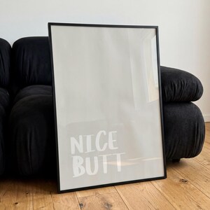 Op de afbeelding: Een ingelijste poster met de woorden "NICE BUTT" in het wit, gecentreerd op een lichtbeige achtergrond. De lijst is zwart en rechthoekig. De poster leunt tegen een donkergrijze, afgeronde bank.