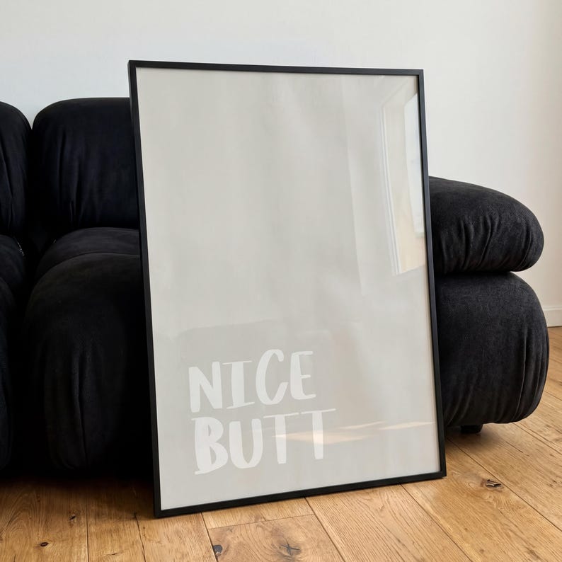 Op de afbeelding: Een ingelijste poster met de woorden "NICE BUTT" in het wit, gecentreerd op een lichtbeige achtergrond. De lijst is zwart en rechthoekig. De poster leunt tegen een donkergrijze, afgeronde bank.