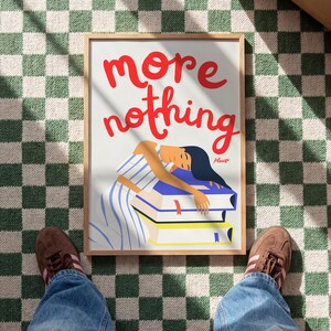 Könnte beinhalten: Ein gerahmter Kunstdruck mit den Worten "more nothing" in roter Schrift. Die Illustration zeigt eine Person, die sich auf einem Stapel Bücher ausruht. Die Person trägt ein blau-weiß gestreiftes Kleid. Das Wort "please" ist in kleiner Schrift geschrieben.