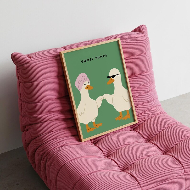 May include: A framed art print featuring two cartoon ducks holding hands against a green background. One duck wears a pink turban, the other sunglasses. The text "GOOSE BUMPS" is at the top. The print is displayed on a pink corduroy chair.
