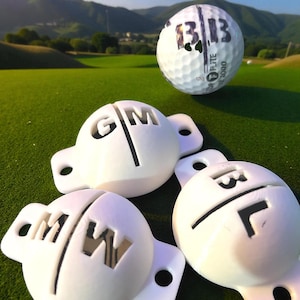 Puede incluir: Una pelota de golf blanca con el número "13" impreso, flotando sobre tres marcadores de pelota blancos. Los marcadores tienen las letras "G", "M" y "W" recortadas. La escena está ambientada en un campo de golf verde.