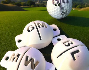 May include: A golf ball with the number "13" printed on it, hovering above three white golf ball markers. The markers have the letters "G", "M", and "W" cut out. The scene is set on a green golf course.