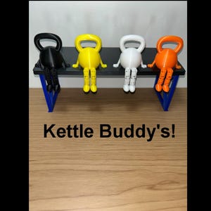 May include: Four colourful kettlebell-shaped figurines with arms and legs, sitting on a black bench with blue supports. The figurines are black, yellow, white, and orange. The text "Kettle Buddy's!" is at the bottom.