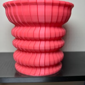 May include: A vibrant coral-colored vase with a unique, layered design. The vase features a series of stacked, rounded sections, each adorned with vertical ridges. The textured surface and bright hue make it a striking decorative piece.