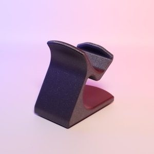 May include: A dark grey, textured phone stand with a curved design. The stand has a rectangular base and a cradle to hold a phone. The interior of the stand is a deep red colour.