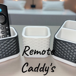 Puede incluir: Tres organizadores de mandos a distancia. Los organizadores son blancos y negros. Los organizadores tienen un patrón geométrico. El texto "Remote Caddy's" está escrito en cursiva negra. Un mando a distancia negro está en uno de los organizadores.