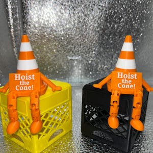 May include: Two novelty figures, each featuring an orange and white traffic cone head, orange arms and legs, and a sign that reads "Hoist the Cone!". One figure sits on a yellow crate, the other on a black crate.