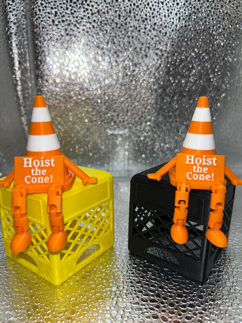 May include: Two novelty figures, each featuring an orange and white traffic cone head, orange arms and legs, and a sign that reads "Hoist the Cone!". One figure sits on a yellow crate, the other on a black crate.
