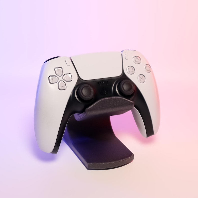 May include: A white and black PlayStation 5 controller on a black stand. The controller has a symmetrical design with buttons and joysticks. The background has a pink and purple gradient.