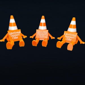 May include: Three orange and white traffic cone figures with articulated arms and legs. Each figure holds a rectangular orange sign with the white text Emotional Support Cone printed on the front. The figures are seated in a row against a dark background.