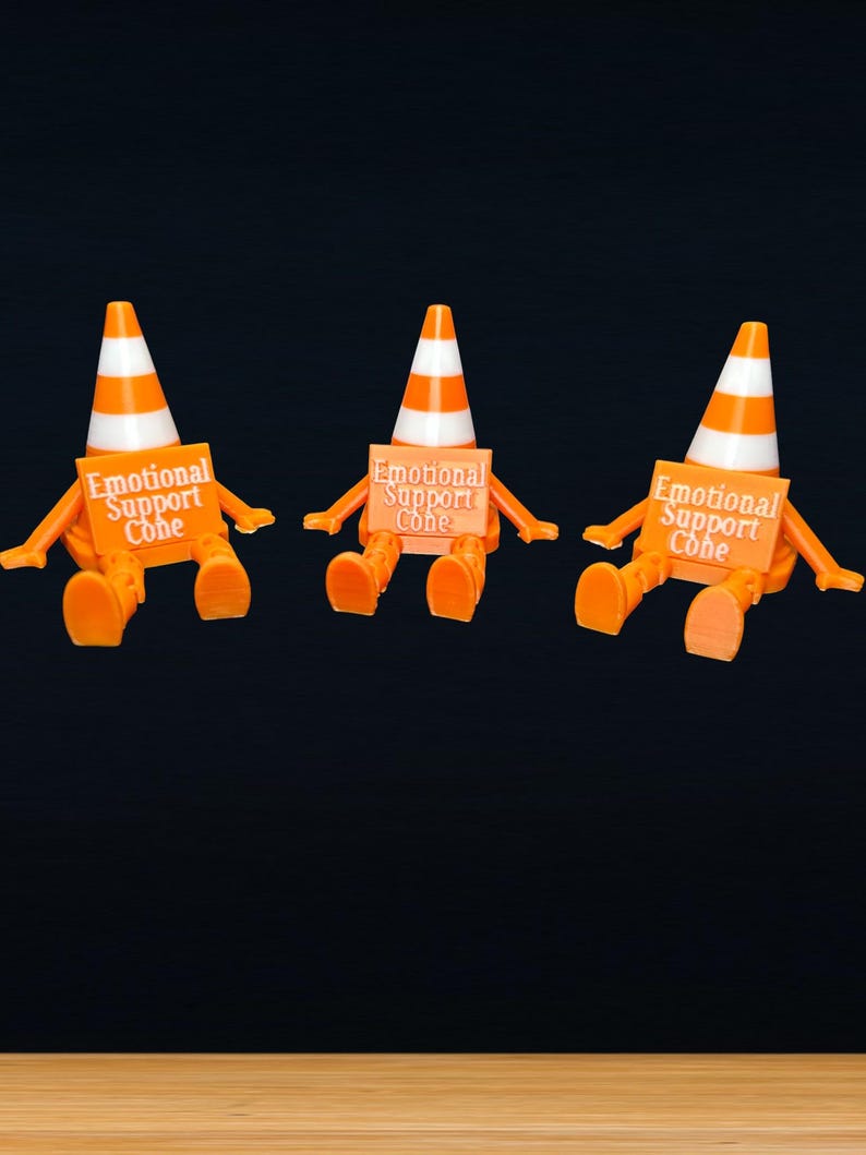 May include: Three orange and white traffic cone figures with articulated arms and legs. Each figure holds a rectangular orange sign with the white text Emotional Support Cone printed on the front. The figures are seated in a row against a dark background.