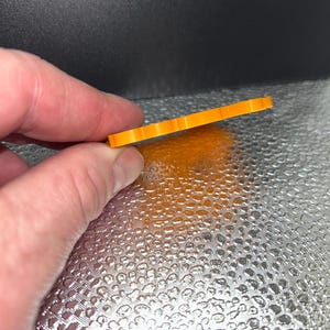 May include: A small, orange, rectangular object with a ridged edge is held between fingers. The object appears to be made of plastic or a similar material. The background is a textured, reflective surface.
