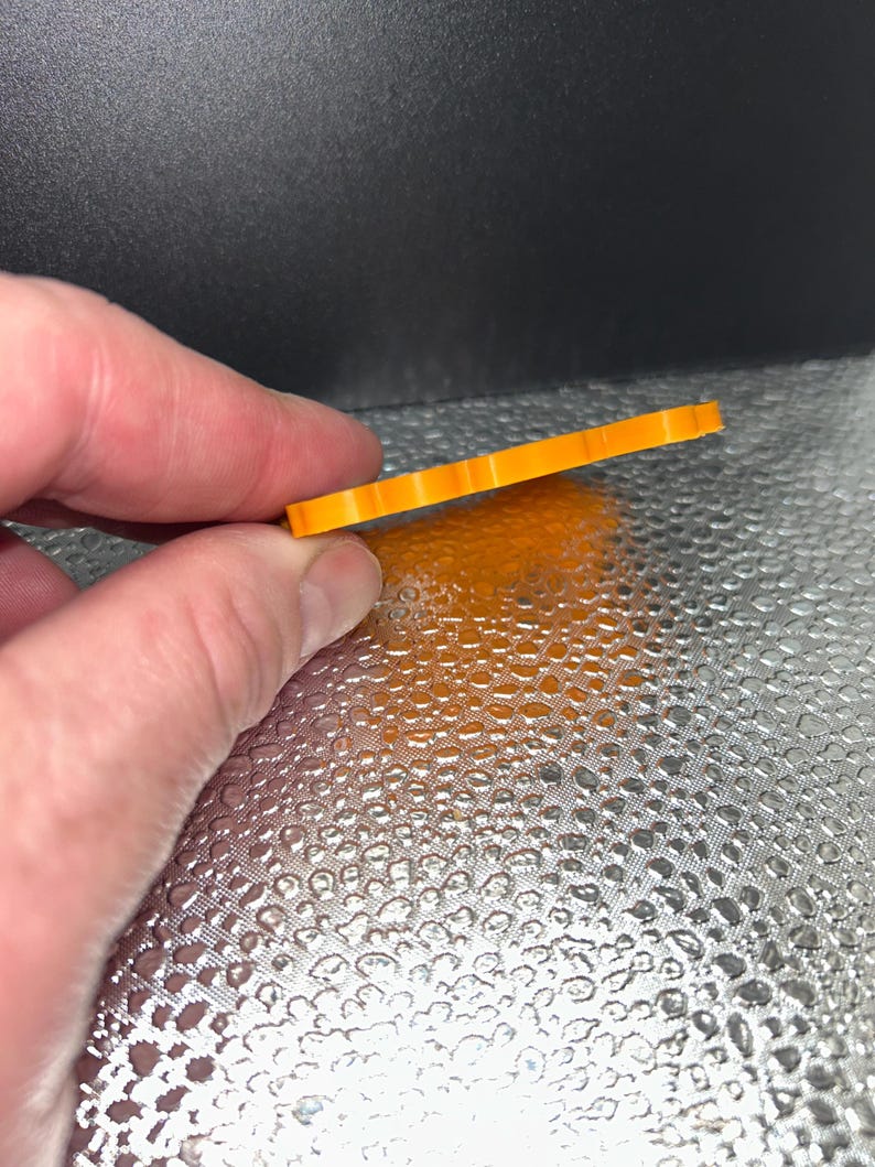 May include: A small, orange, rectangular object with a ridged edge is held between fingers. The object appears to be made of plastic or a similar material. The background is a textured, reflective surface.