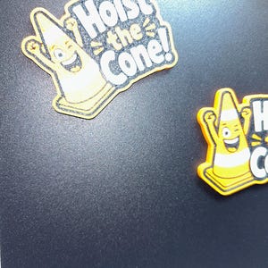 May include: Two yellow and orange magnets with the text "Hoist the Cone!" are displayed. The magnets feature a smiling cartoon traffic cone character with raised arms. The text is in white with a black outline.