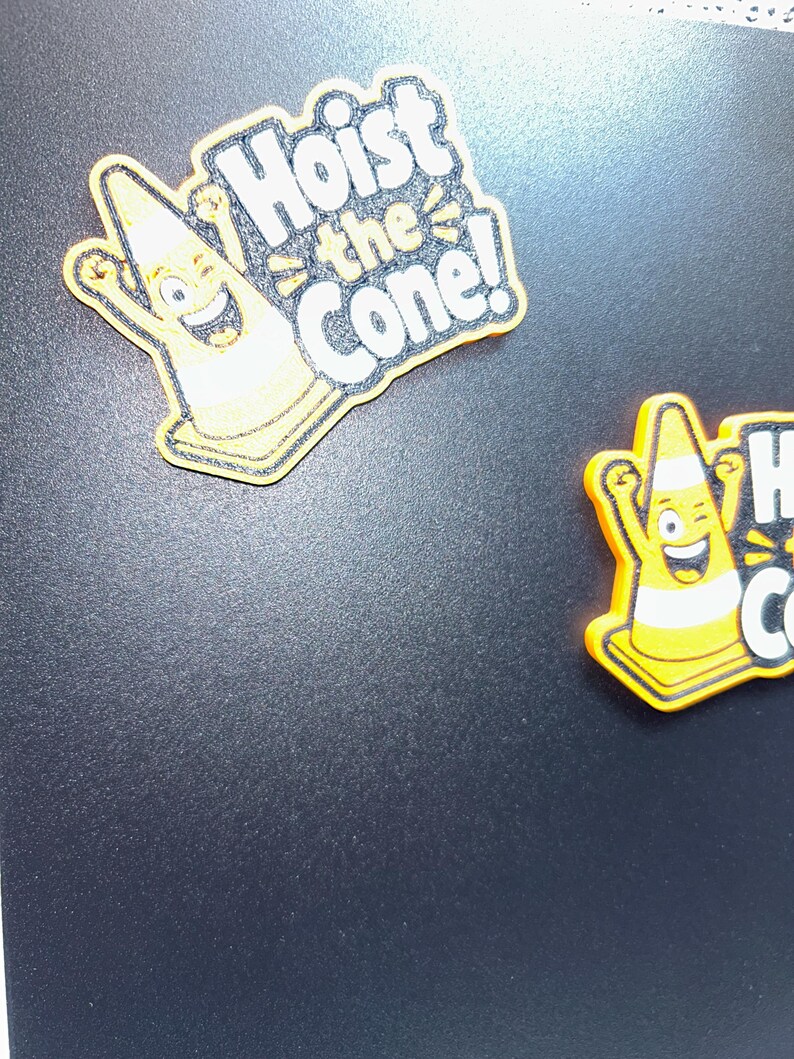 May include: Two yellow and orange magnets with the text "Hoist the Cone!" are displayed. The magnets feature a smiling cartoon traffic cone character with raised arms. The text is in white with a black outline.