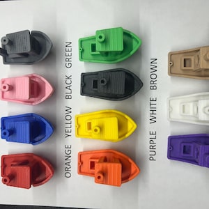May include: A selection of small, 3D-printed toy boats in a range of colours: grey, pink, blue, red, green, black, yellow, orange, brown, white, and purple. Each boat features a cabin and a small smokestack.