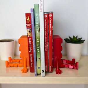 May include: A collection of books and video game cases held between orange and red decorative bookends. The books include titles like "Mothers & Daughters" and "For the Love of Dogs". The bookends have a unique, playful design.