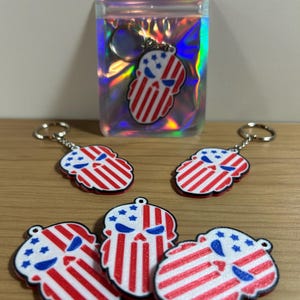May include: Keychains featuring a skull design with a patriotic American flag theme. The keychains are red, white, and blue, with a skull shape and a metal keyring. One keychain is inside a holographic bag.