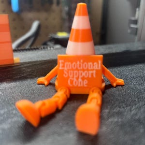 Pode incluir: Uma figura de cone de trânsito laranja e branco com braços e pernas. A figura tem as palavras "Emotional Support Cone" impressas em seu corpo. O cone está sentado em uma superfície escura.