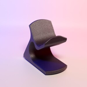 May include: A black stand with a curved design and a textured top. The stand has a rounded base and a curved support. The background fades from purple to pink.