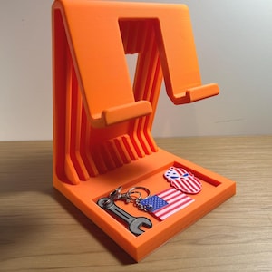 May include: Vibrant orange 3D-printed phone stand with a tray. The stand features a phone slot and a small tray holding a wrench keyring, a miniature American flag, and a patriotic pin.