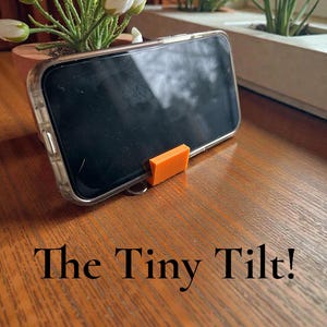 May include: A smartphone in a clear case is propped up by a small orange stand. The phone rests on a wooden surface, with the text "The Tiny Tilt!" below. Decorative plants are in the background.