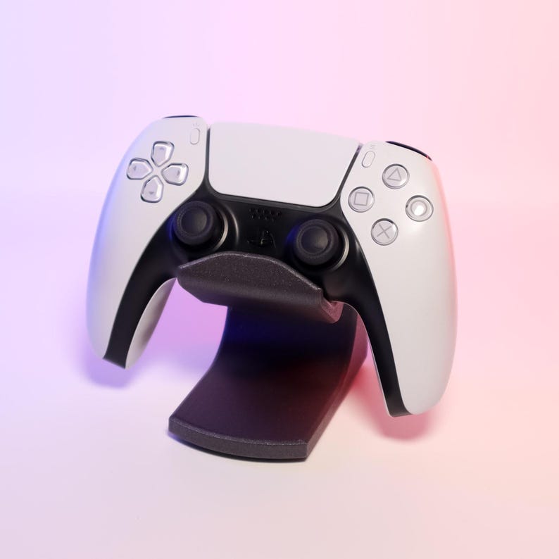May include: A white and black games controller on a black stand. The controller features a symmetrical design with buttons and joysticks. The stand is a curved shape, designed to hold the controller upright.