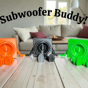 May include: Three colorful subwoofer speaker stands in orange, gray, and green, each with a speaker design and legs. The text "Subwoofer Buddy!" is arched above the speakers. The stands are on a wooden surface.