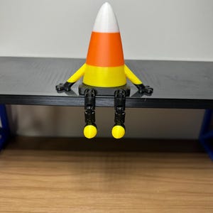 Peut inclure: Une figurine de bonbon en forme de maïs, imprimée en 3D, avec des bras et des jambes, assise sur une surface noire. Le bonbon a une pointe blanche, un milieu orange et une base jaune. Les bras sont jaunes avec des mains noires et les jambes sont noires avec des pieds jaunes.