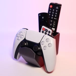 K&ouml;nnte beinhalten: Ein wei&szlig;-schwarzer PlayStation 5-Controller liegt auf einem grauen St&auml;nder, mit einem schwarzen Halter, der drei Fernbedienungen enth&auml;lt. Die Fernbedienungen sind schwarz mit wei&szlig;en Tasten, und eine kleine Fernbedienung hat den Text "ON OFF".