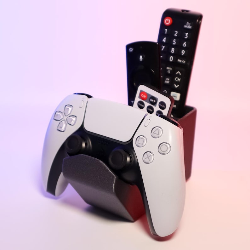 K&ouml;nnte beinhalten: Ein wei&szlig;-schwarzer PlayStation 5-Controller liegt auf einem grauen St&auml;nder, mit einem schwarzen Halter, der drei Fernbedienungen enth&auml;lt. Die Fernbedienungen sind schwarz mit wei&szlig;en Tasten, und eine kleine Fernbedienung hat den Text "ON OFF".