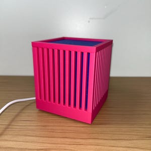 May include: A vibrant pink cube-shaped lamp with vertical bar detailing. The interior is a deep blue, and a white cord extends from the base. The lamp sits on a wooden surface, offering a modern, geometric design.