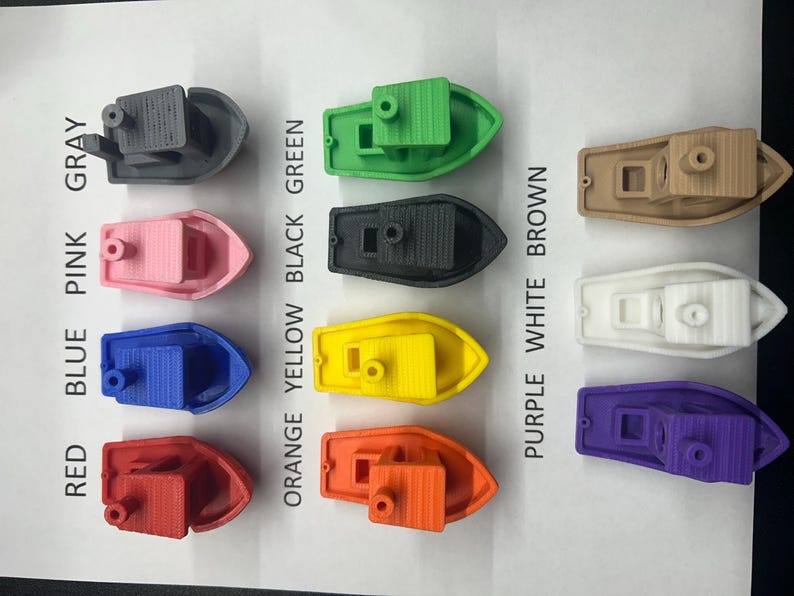 K&ouml;nnte beinhalten: Eine Sammlung kleiner, 3D-gedruckter Spielzeugboote in verschiedenen Farben, darunter Grau, Rosa, Blau, Rot, Gr&uuml;n, Schwarz, Gelb, Orange, Braun, Wei&szlig; und Lila. Jedes Boot hat eine Kabine und einen kleinen Schornstein.