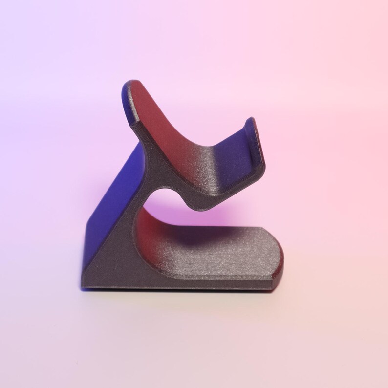 May include: A dark grey phone stand with a curved design. The stand features a base, a curved support, and a cradle for a phone. It's made of a textured material and set against a pink and blue gradient background.