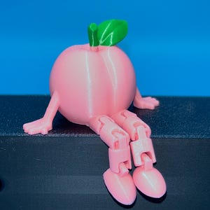 May include: A pink 3D-printed peach figurine with articulated arms and legs. The peach has a green leaf and stem. The figurine is sitting on a black surface against a blue background.