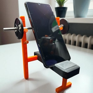 Peut inclure: Un support de téléphone orange et noir imprimé en 3D en forme de banc de musculation. Il maintient un smartphone noir, avec des poids d'haltères de chaque côté. Le support est sur une surface blanche.
