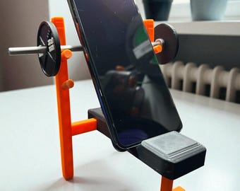 May include: A 3D-printed orange and black phone stand designed as a weightlifting bench. It holds a black smartphone, with barbell weights on either side. The stand is on a white surface.