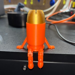 Könnte beinhalten: Eine 3D-gedruckte orangefarbene und goldfarbene Figur, die einer Kugel mit Armen und Beinen ähnelt. Die Figur sitzt auf einer schwarzen Oberfläche, mit einem Werkstatt-Hintergrund.