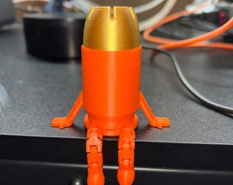 May include: A 3D-printed orange and gold figurine, shaped like a bullet, with arms and legs. The figure is seated on a black surface, with a workshop background visible.