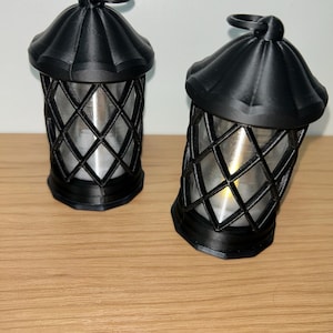 Pode incluir: Duas lanternas decorativas pretas com um invólucro de vidro com padrão de diamante e uma pequena alça na parte superior. As lanternas têm uma luz semelhante a uma vela no interior, criando um brilho quente. Estão sobre uma superfície de madeira.