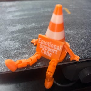 May include: A small orange and white traffic cone figurine with articulated orange arms and legs. The cone features a rectangular orange sign attached to the front with the white text "Emotional Support Cone" printed clearly on the surface.
