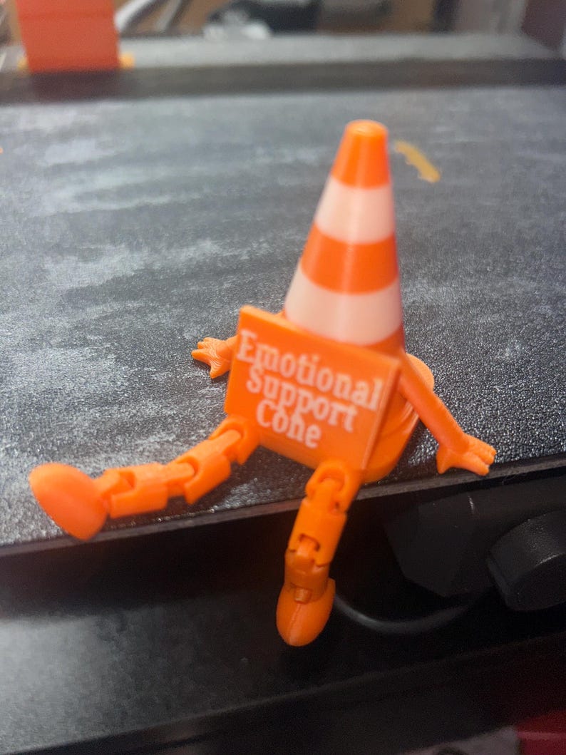 May include: A small orange and white traffic cone figurine with articulated orange arms and legs. The cone features a rectangular orange sign attached to the front with the white text "Emotional Support Cone" printed clearly on the surface.
