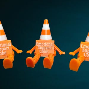 May include: Three orange and white traffic cone figures with articulated limbs, each holding a rectangular orange sign that reads Emotional Support Cone in white raised lettering. The figures are seated in a row against a dark background.