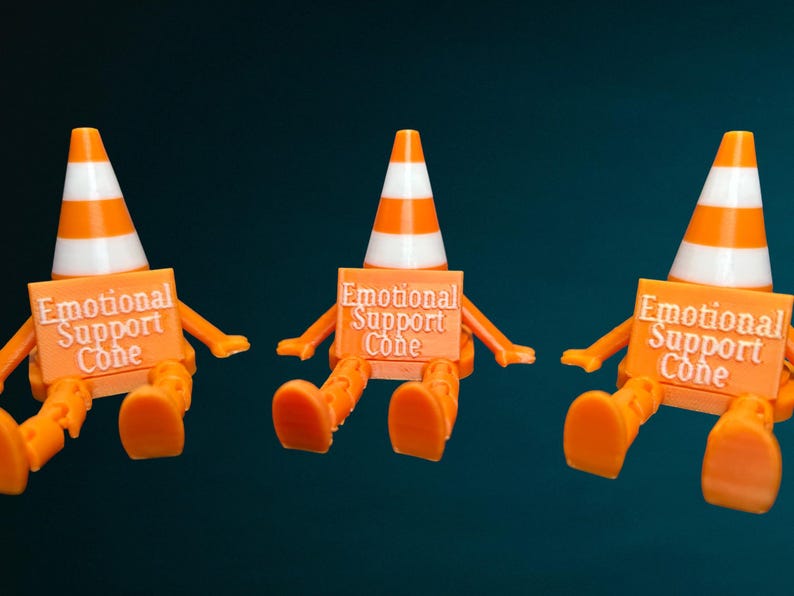 May include: Three orange and white traffic cone figures with articulated limbs, each holding a rectangular orange sign that reads Emotional Support Cone in white raised lettering. The figures are seated in a row against a dark background.