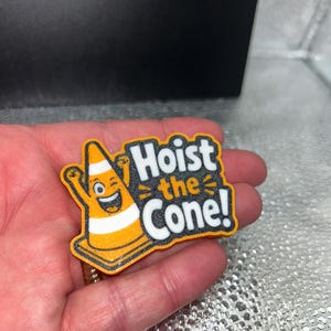 May include: A colorful, cartoonish pin featuring a smiling traffic cone with arms raised. The text "Hoist the Cone!" is displayed in bold, white letters with a black outline, set against an orange and black background.