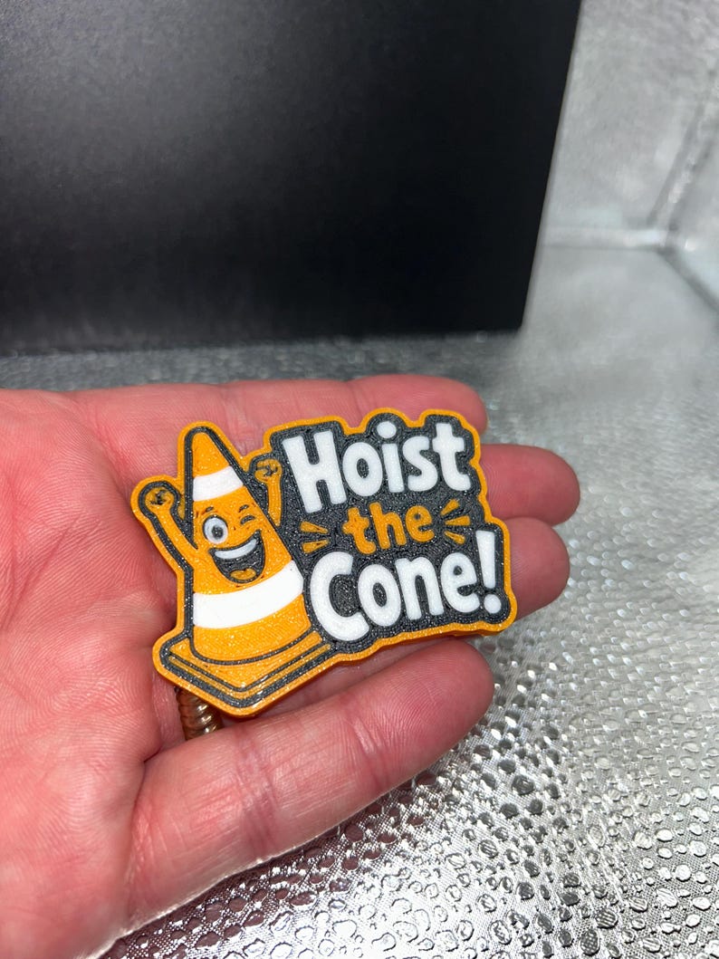 May include: A colorful, cartoonish pin featuring a smiling traffic cone with arms raised. The text "Hoist the Cone!" is displayed in bold, white letters with a black outline, set against an orange and black background.