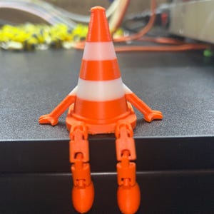 Può includere: Una statuina stampata in 3D a forma di cono stradale arancione e bianco con gambe e braccia articolate. Il cono presenta strisce bianche ed è posizionato su una superficie scura. Un'interpretazione giocosa di un comune oggetto di sicurezza stradale.