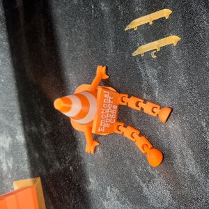 May include: An orange and white traffic cone figure with articulated limbs and a square sign reading Emotional Support Cone. The figure has two arms and two legs with rounded feet. Two separate gold-colored blade-like accessories are included.